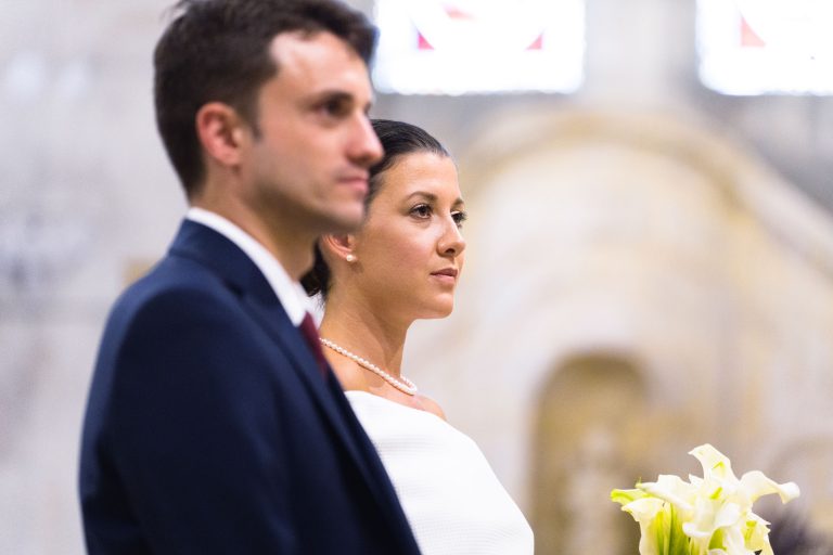 Une mariée, Valentine et un marié se tiennent côte à côte à l'intérieur, de face. La mariée porte une robe blanche et tient des lys calla ; l'arrière-plan est légèrement flou, suggérant un décor orné à la mairie de Camblanes-et-Meynac.