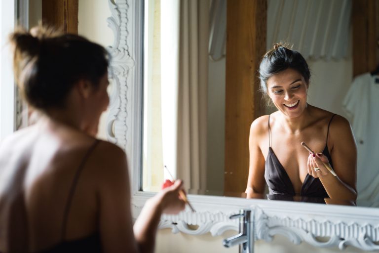 Une femme aux cheveux bruns coiffés en chignon sourit tout en se maquillant au pinceau, debout devant un miroir décoratif à cadre blanc. Se préparant pour la Saint-Valentin au Château de Garde à Moulon, la mariée porte un haut sombre sans manches alors que la lumière naturelle remplit la pièce.