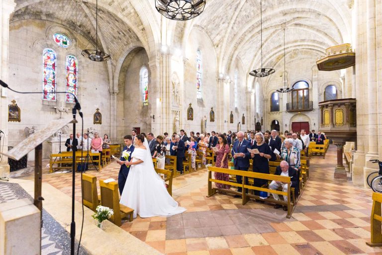 Les mariés descendent l'allée dans une grande église en pierre, qui rappelle l'élégance du Château de Garde à Moulon, avec ses hauts plafonds voûtés et ses vitraux. Les invités en tenue de soirée les observent depuis les bancs en bois chaleureusement éclairés.
