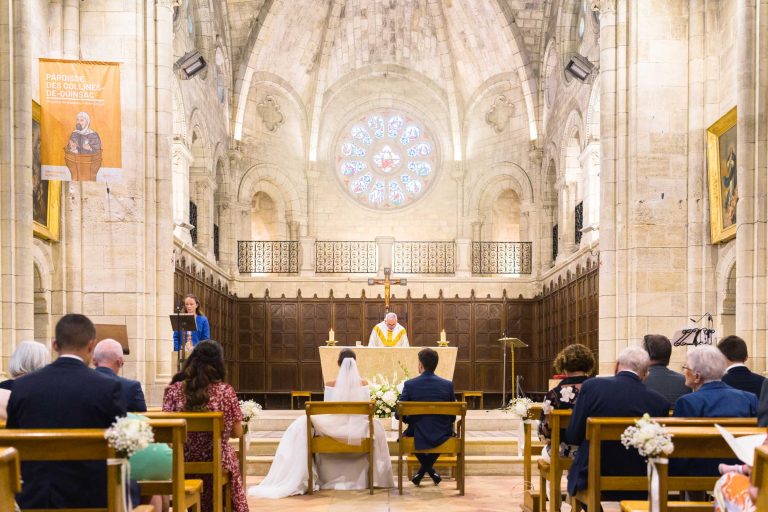 Cérémonie de mariage à l'intérieur d'une grande église en pierre. La mariée et le marié sont assis face à l'autel, où se tient un prêtre en robe blanche et or. Les invités sont assis sur des bancs. Une grande rosace, de hautes arches et des bannières décorent l'intérieur lumineux.