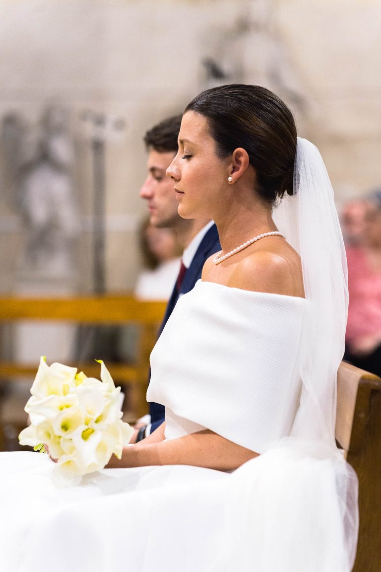 Une mariée vêtue d'une robe blanche à épaulettes et d'un voile est assise à l'intérieur, tenant un bouquet de lys calla blancs. Valentine porte un collier de perles et semble sereine, les yeux fermés. Le marié, Antonin, en costume sombre, est assis à côté d'elle, l'arrière-plan étant légèrement flou.