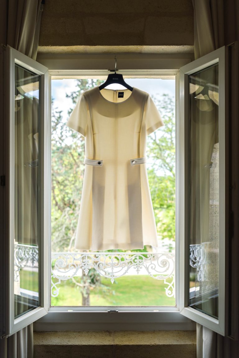 Une robe à manches courtes de couleur crème est suspendue à un cintre noir dans une fenêtre ouverte. La lumière du soleil filtre à travers la robe, parfaite pour la mariée, avec des arbres verdoyants à l'extérieur et une délicate ferronnerie décorative sous la fenêtre à cadre blanc.