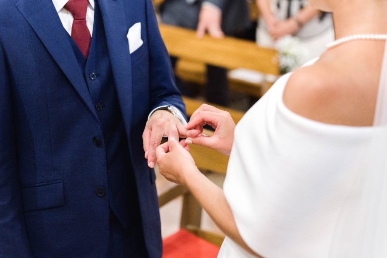 Une mariée en robe blanche glisse une alliance au doigt du marié. Valentine et son époux, Antonin, se tiennent l'un près de l'autre dans un décor intérieur lumineux, avec des bancs en bois et des invités flous à l'arrière-plan.