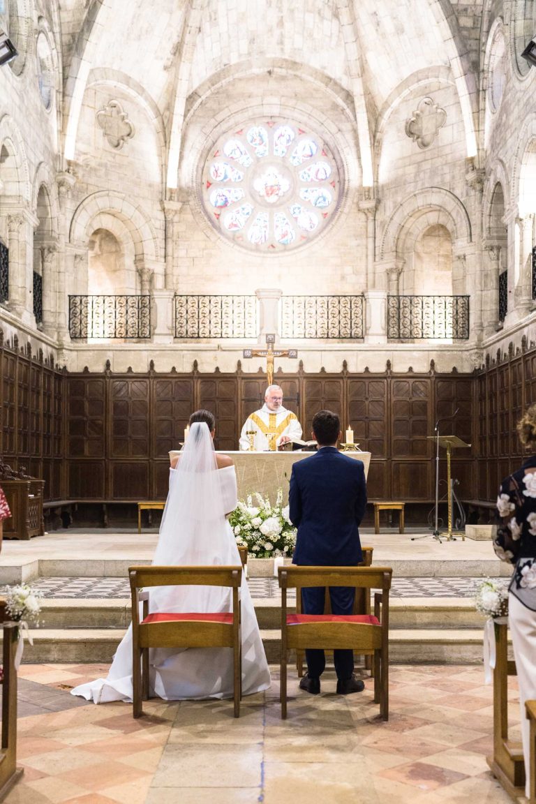 La mariée et le marié sont agenouillés devant l'autel d'une grande église en pierre. Le prêtre se tient devant eux, les mains levées, sous un grand vitrail orné. Le plafond voûté et les fleurs créent une atmosphère solennelle et respectueuse pour le jour spécial de la Saint-Valentin.