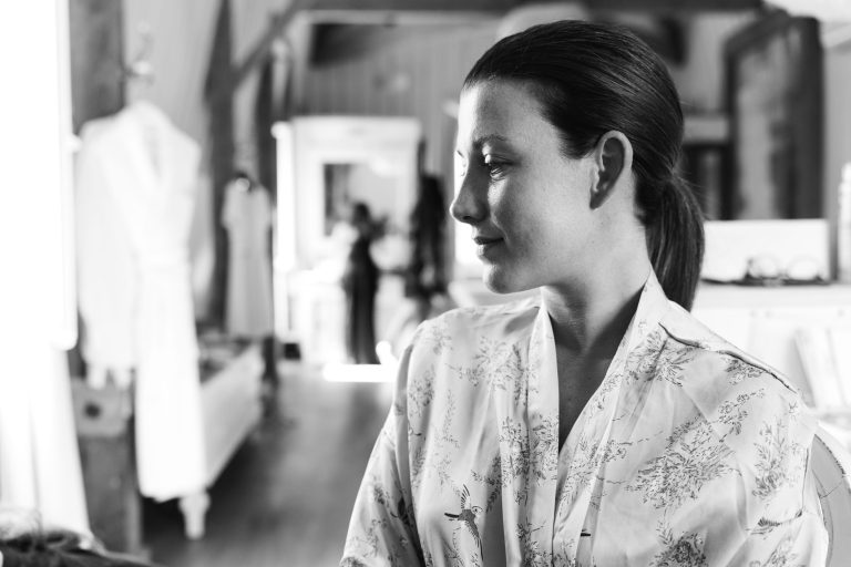 Une femme aux cheveux noirs en queue de cheval, vêtue d'une robe à fleurs, se tient à l'intérieur et regarde vers la droite. L'image capture la mariée en noir et blanc, dans une salle doucement éclairée du château de Garde à Moulon.