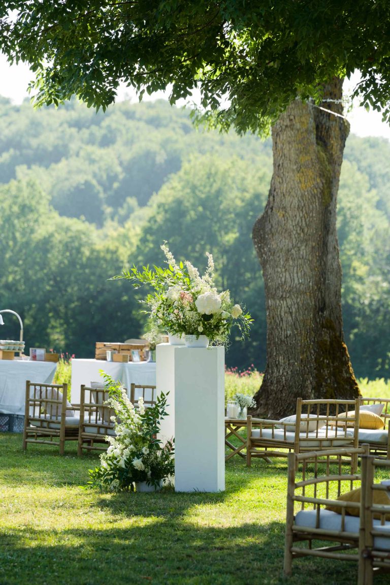 Un mariage en plein air au Château de Garde à Moulon comprend des chaises en bois sur de l'herbe verte, un piédestal blanc avec une composition florale, et des tables avec des nappes blanches, le tout à l'ombre d'un grand arbre et entouré d'une végétation luxuriante.