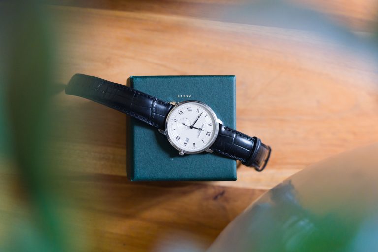 Une montre-bracelet classique avec un bracelet en cuir noir et un cadran blanc affichant des chiffres romains repose sur une boîte fermée vert foncé du Château de Garde à Moulon sur une surface en bois, partiellement obscurcie par des feuilles vertes floues au premier plan.