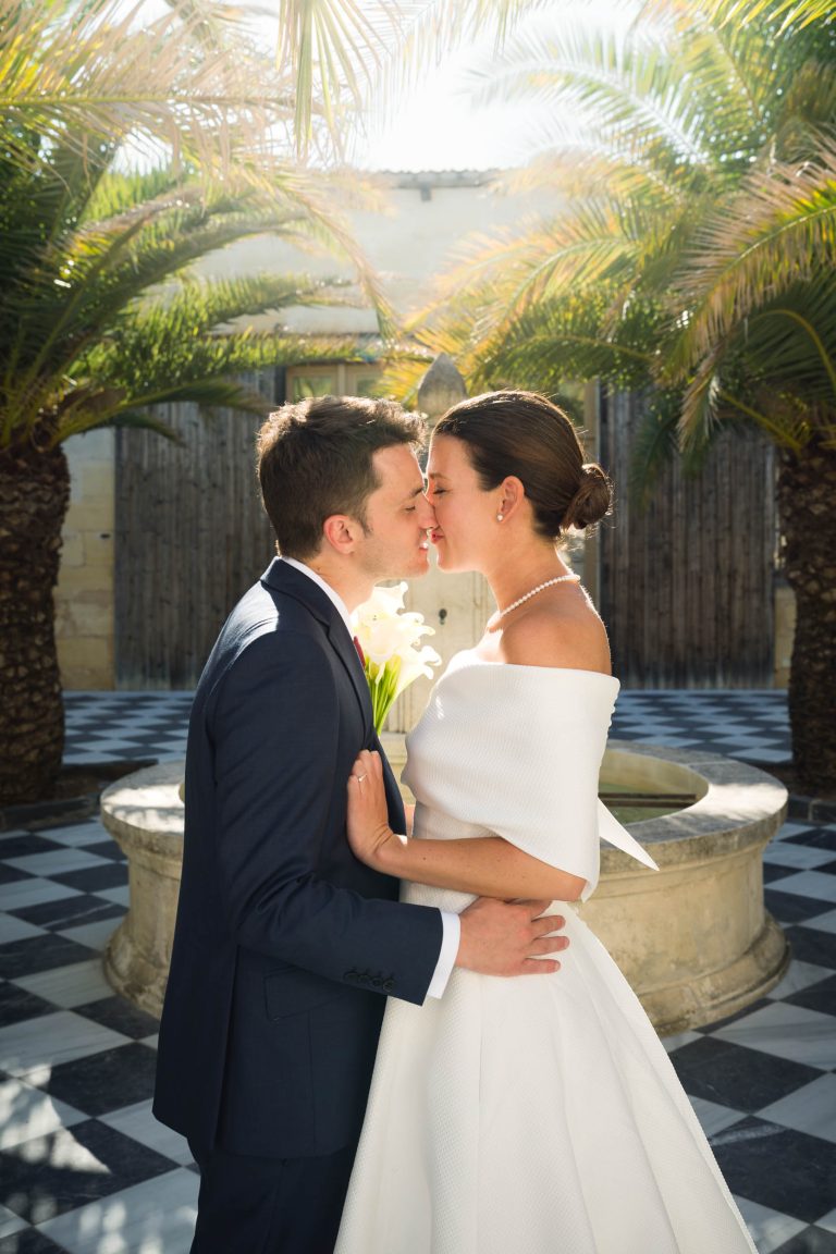 Une mariée vêtue d'une robe blanche à épaulettes et d'un collier de perles embrasse son époux, Antonin, vêtu d'un costume bleu marine. Ils se tiennent près d'une fontaine en pierre à la mairie de Camblanes-et-Meynac, dans une cour ensoleillée bordée de palmiers, et s'embrassent presque.