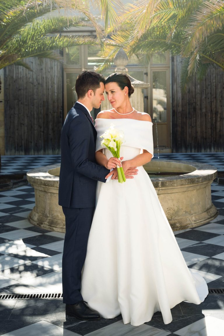 Une mariée vêtue d'une robe blanche à épaulettes et tenant des lys calla se tient près du marié vêtu d'un costume bleu marine. Ils se sourient doucement, se touchant le front sur un sol à carreaux près d'une fontaine en pierre au Château de Garde à Moulon, encadrés par des palmiers et la lumière du soleil.