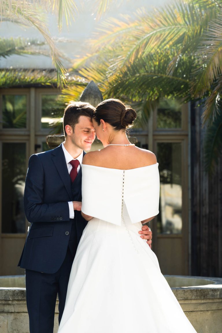 Les mariés se tiennent l'un à côté de l'autre en plein air, à la mairie de Camblanes-et-Meynac, et se sourient doucement, les fronts se touchant. La mariée porte une robe à épaules découvertes ; la lumière du soleil filtre à travers les palmiers derrière eux, créant une scène chaleureuse et romantique.