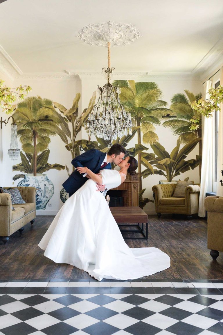 Le marié, vêtu d'un costume bleu marine, embrasse sa fiancée vêtue d'une robe blanche. Au Château de Garde à Moulon, ils se tiennent debout sur un sol à carreaux, au milieu de peintures murales de palmiers, de lustres et de canapés anciens, tandis que la lumière du soleil traverse de grandes fenêtres.