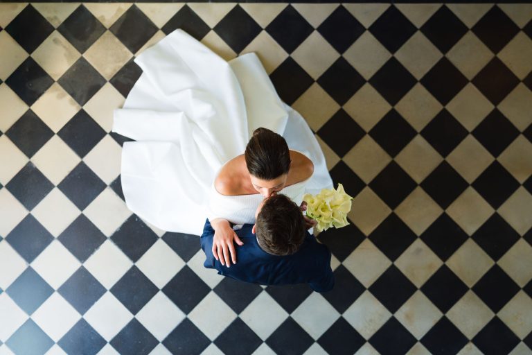 Une mariée en robe blanche et le marié en costume sombre s'embrassent sur un sol à carreaux noirs et blancs. Photographiée de haut à la mairie de Camblanes-et-Meynac, la mariée tient des fleurs jaunes tandis qu'ils partagent un moment romantique, semblable à une danse.