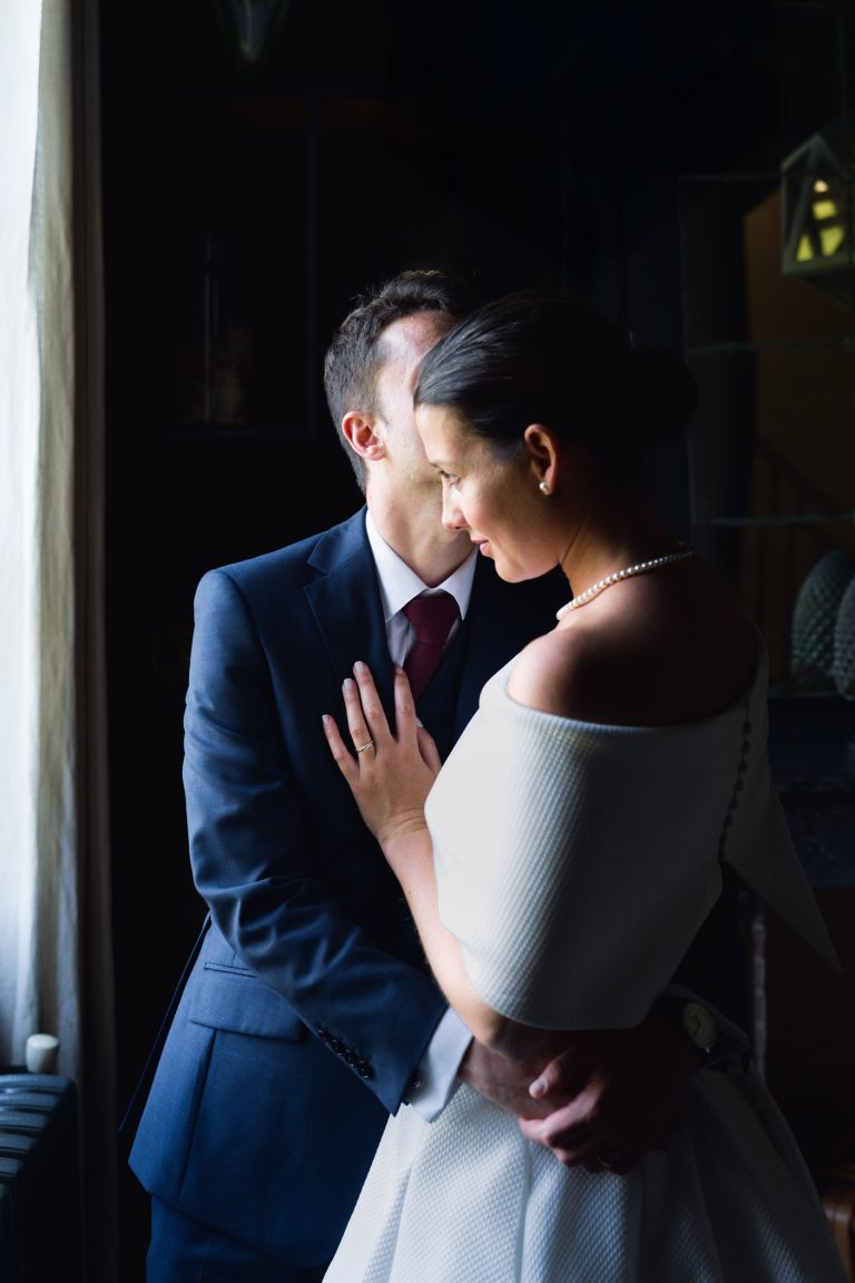 La mariée, vêtue d'une robe blanche décolletée et d'un collier de perles, embrasse le marié, vêtu d'un costume sombre et d'une cravate rouge, près d'une fenêtre, doucement éclairée par la lumière naturelle. C'est un moment tendre de la Saint-Valentin, où leurs visages se rapprochent doucement l'un de l'autre.