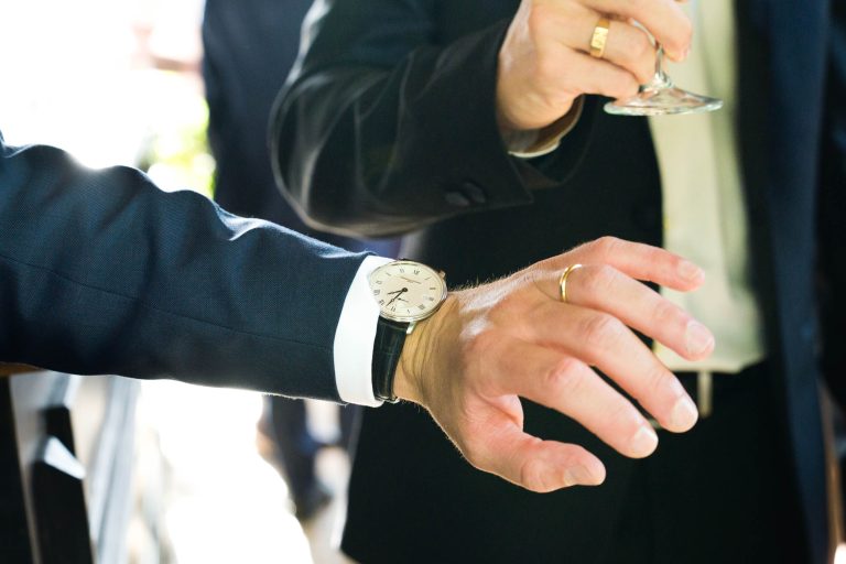 Deux personnes en costume interagissent étroitement ; l'une d'elles, peut-être le marié, porte une alliance en or et une montre-bracelet en argent à 5:40. L'autre tient un verre à vin avec une bague en or et des boutons de manchette. Un arrière-plan flou suggère un événement formel de la Saint-Valentin.