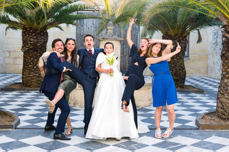 Six personnes posent de manière ludique lors d'un mariage ; la mariée en blanc tient des fleurs, entourée d'invités qui font des grimaces et des gestes dramatiques sur un sol à carreaux noirs et blancs, avec des palmiers et un mur de pierre en arrière-plan.