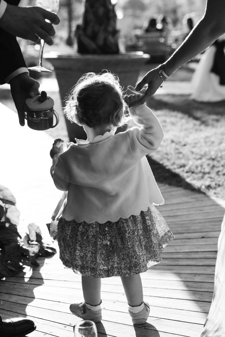 Un bambin aux cheveux bouclés, vêtu d'une robe et d'un gilet à fleurs, marche main dans la main avec la mariée le long d'un chemin de bois éclairé par le soleil. Un autre adulte lui offre une tasse à bec, tandis que les gens de la mairie de Camblanes-et-Meynac sont légèrement flous à l'arrière-plan.