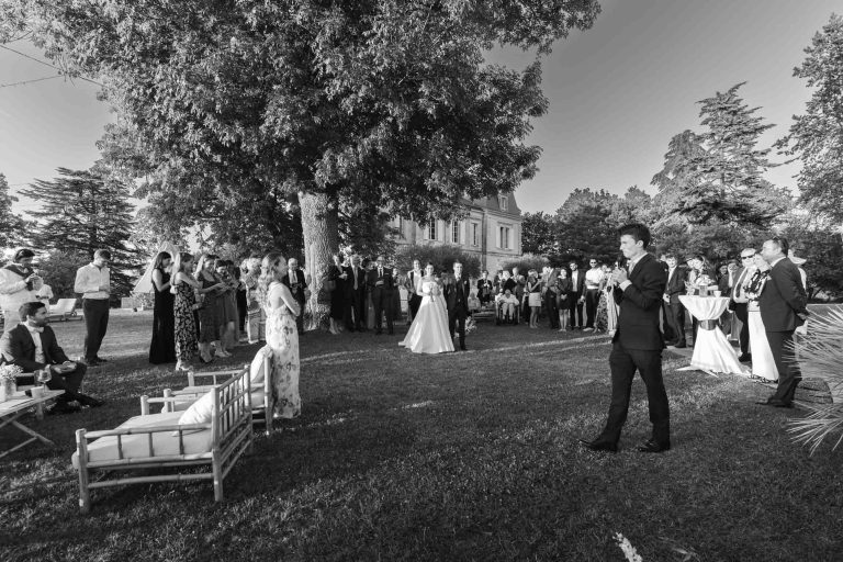Un grand groupe en tenue de soirée se réunit en plein air sur la pelouse du Château de Garde à Moulon. Valentine et Antonin se tiennent au centre, sous le regard des invités, un homme tenant un micro - peut-être en train de prononcer un discours animé et festif.