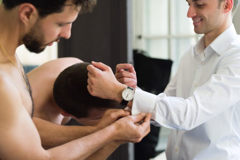 Trois hommes se préparent pour un événement formel. Antonin, en chemise blanche, sourit tandis qu'un autre homme, torse nu, l'aide à ajuster sa manchette. Valentine l'assiste, concentrée sur la montre et la manchette. La scène se déroule à l'intérieur, avec une lumière naturelle à travers la fenêtre.