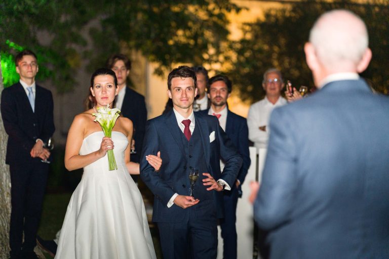 La mariée en robe blanche sans bretelles tient des fleurs au bras du marié en costume bleu marine. Ils avancent ensemble vers un homme chauve, lors d'un événement du soir devant la mairie de Camblanes-et-Meynac.