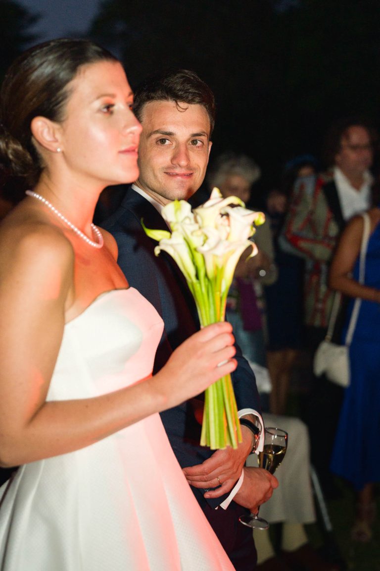 Une mariée en robe blanche sans bretelles tient des lys calla, face à la gauche. Le marié, souriant dans un costume sombre, se tient derrière elle avec un verre de vin. Les invités de Valentine, en tenue colorée, se fondent dans l'arrière-plan du Château de Garde à Moulon.