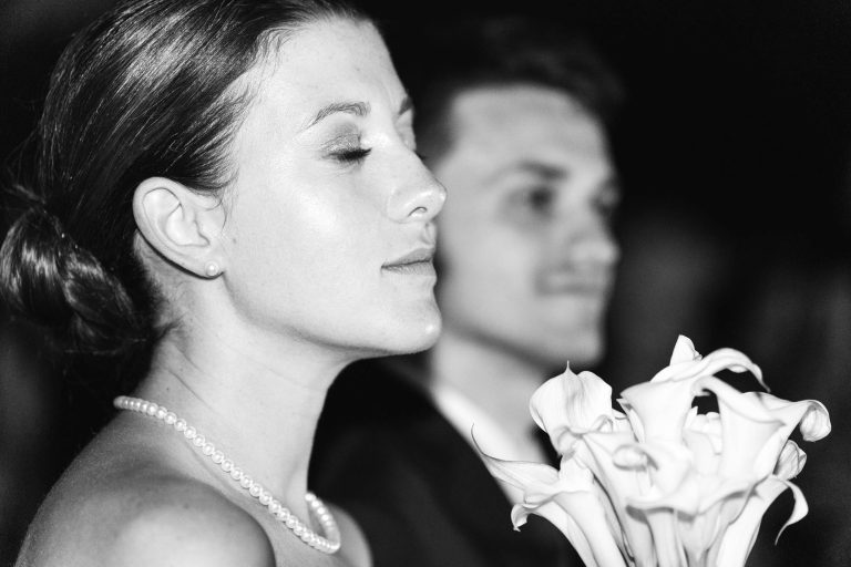 Une femme aux yeux fermés tient un bouquet de callas blancs au Château de Garde à Moulon. Elle porte un collier de perles, les cheveux en chignon. À ses côtés, un homme en costume est légèrement flou ; l'image en noir et blanc respire la sérénité.