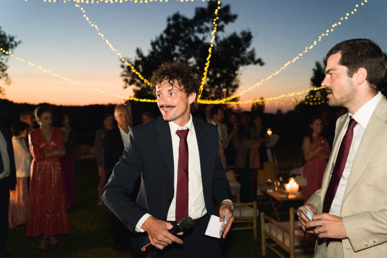 Un homme en costume sombre et cravate rouge tient un microphone et des fiches, souriant lors d'une soirée en plein air avec des guirlandes lumineuses au-dessus de sa tête. À proximité, Antonin se tient debout en costume clair tandis que les invités - et peut-être la mariée - se mélangent dans l'arrière-plan doucement éclairé.