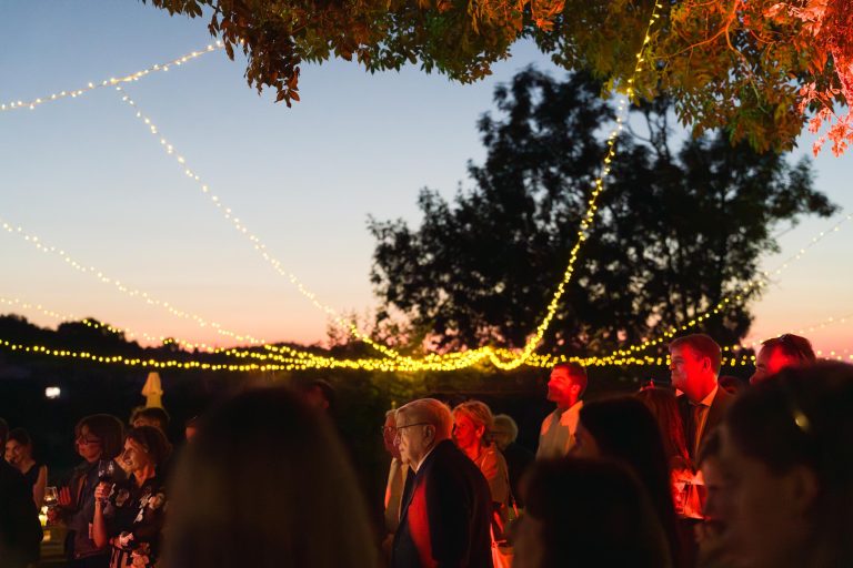 Un groupe de personnes se réunit en plein air au coucher du soleil sous des branches d'arbres et des guirlandes lumineuses près du Château de Garde à Moulon. Le ciel passe de l'orange au bleu profond, et les invités chaleureusement éclairés créent une atmosphère festive et sereine sous la silhouette d'un grand arbre.