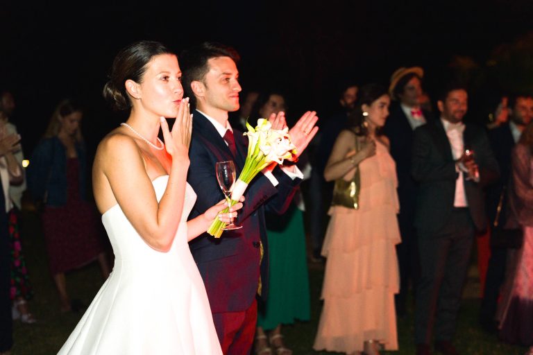 La mariée en robe blanche tient un bouquet et envoie un baiser, aux côtés du marié applaudit. Tous deux font face à leurs invités élégants, baignés de lumière chaleureuse lors d'une nuit magique au Château de Garde à Moulon.