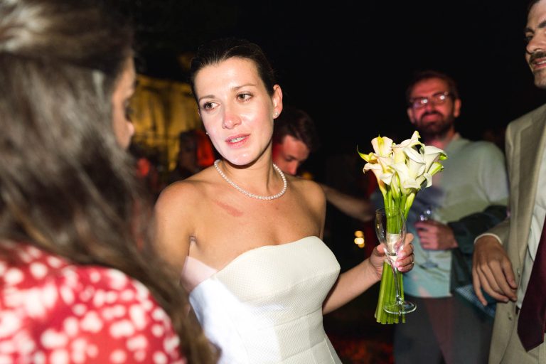 Une mariée en robe blanche sans bretelles et collier de perles tient des lys calla, discutant avec une femme en robe rouge à motifs. Les invités se mêlent à l'arrière-plan du Château de Garde à Moulon lors de la célébration nocturne d'Antonin et du marié.