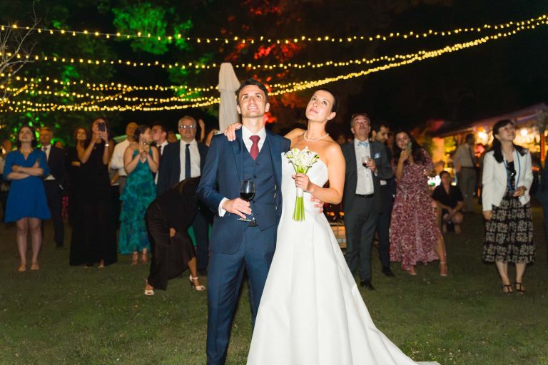 Une mariée en robe blanche tenant des fleurs se tient à côté du marié Antonin en costume bleu marine ; tous deux regardent vers le haut en souriant. Entourés d'invités en plein air, la nuit, sous les guirlandes lumineuses de la mairie de Camblanes-et-Meynac, la scène rayonne de joie.