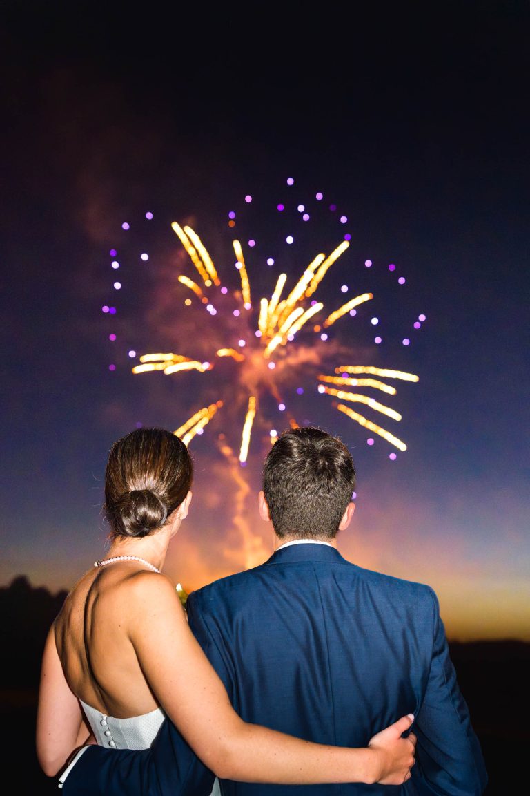 Un couple, dont la mariée en robe bustier et perles, se tient bras dessus bras dessous, dos à l'appareil photo, et regarde un feu d'artifice jaune et violet éclater au-dessus de la mairie de Camblanes-et-Meynac. Le ciel s'illumine tandis qu'ils célèbrent ensemble.