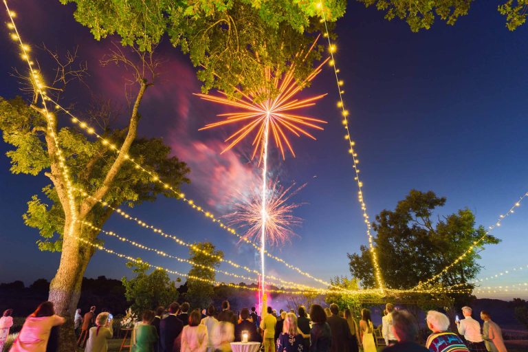 Une foule se rassemble en plein air au crépuscule, sous des guirlandes lumineuses, pour regarder les feux d'artifice rouges et roses de la Saint-Valentin exploser dans le ciel. De grands arbres entourent la scène festive près de la mairie de Camblanes-et-Meynac, alors que le ciel passe du bleu profond aux tons du coucher de soleil.