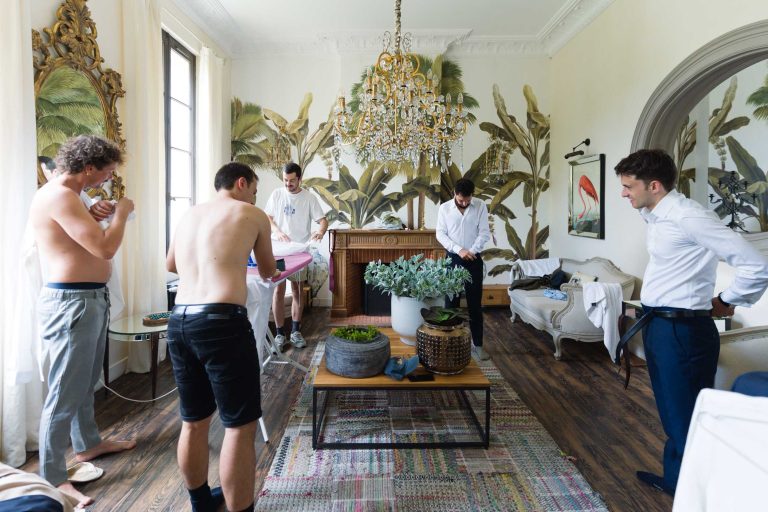 Cinq hommes, dont certains sont torse nu, se préparent dans une pièce élégante du Château de Garde à Moulon. L'un repasse une chemise tandis que le marié se rase, tandis que d'autres s'habillent ou ajustent leurs vêtements parmi des plantes et des tapis à motifs, suggérant des préparatifs avant l'événement.