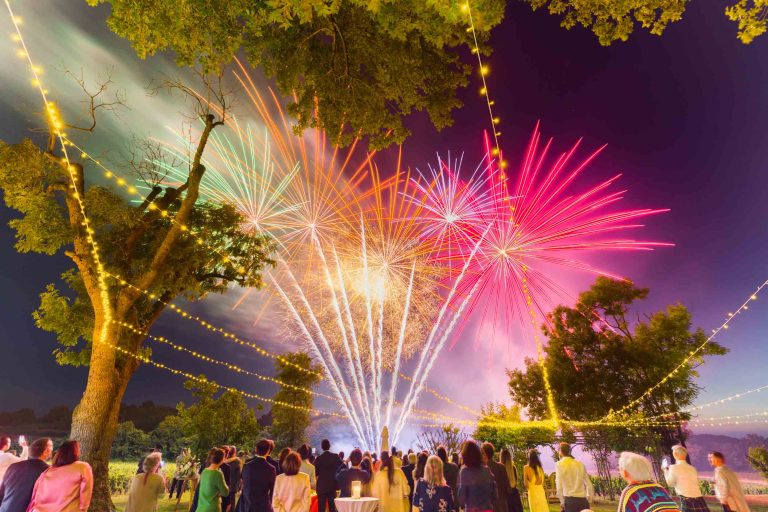 Une foule se rassemble en plein air, la nuit, au Château de Garde à Moulon, pour regarder les feux d'artifice colorés qui éclatent dans le ciel. Des guirlandes lumineuses ajoutent à l'ambiance festive, tandis que la mariée et ses invités admirent les rouges, roses et or éblouissants encadrés par les arbres.