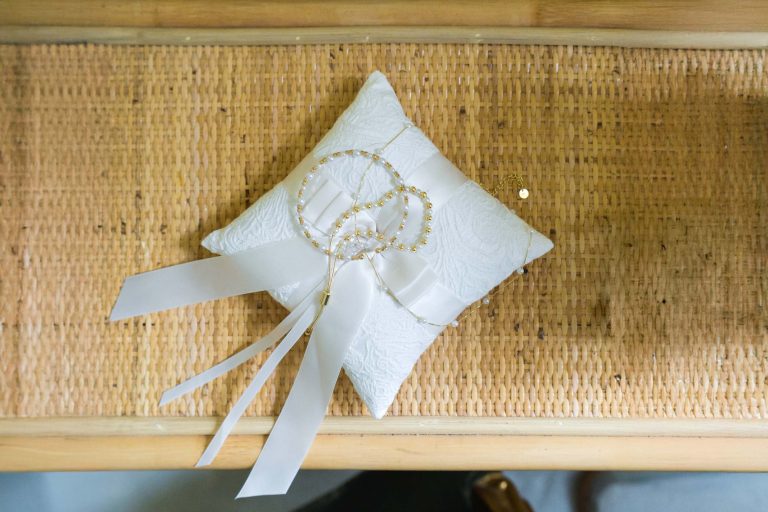 Un coussin de dentelle blanche avec deux couronnes de mariage dorées (stefana) reliées par des rubans blancs repose sur une surface en bois tressé, symbolisant un rituel de mariage orthodoxe grec traditionnel.