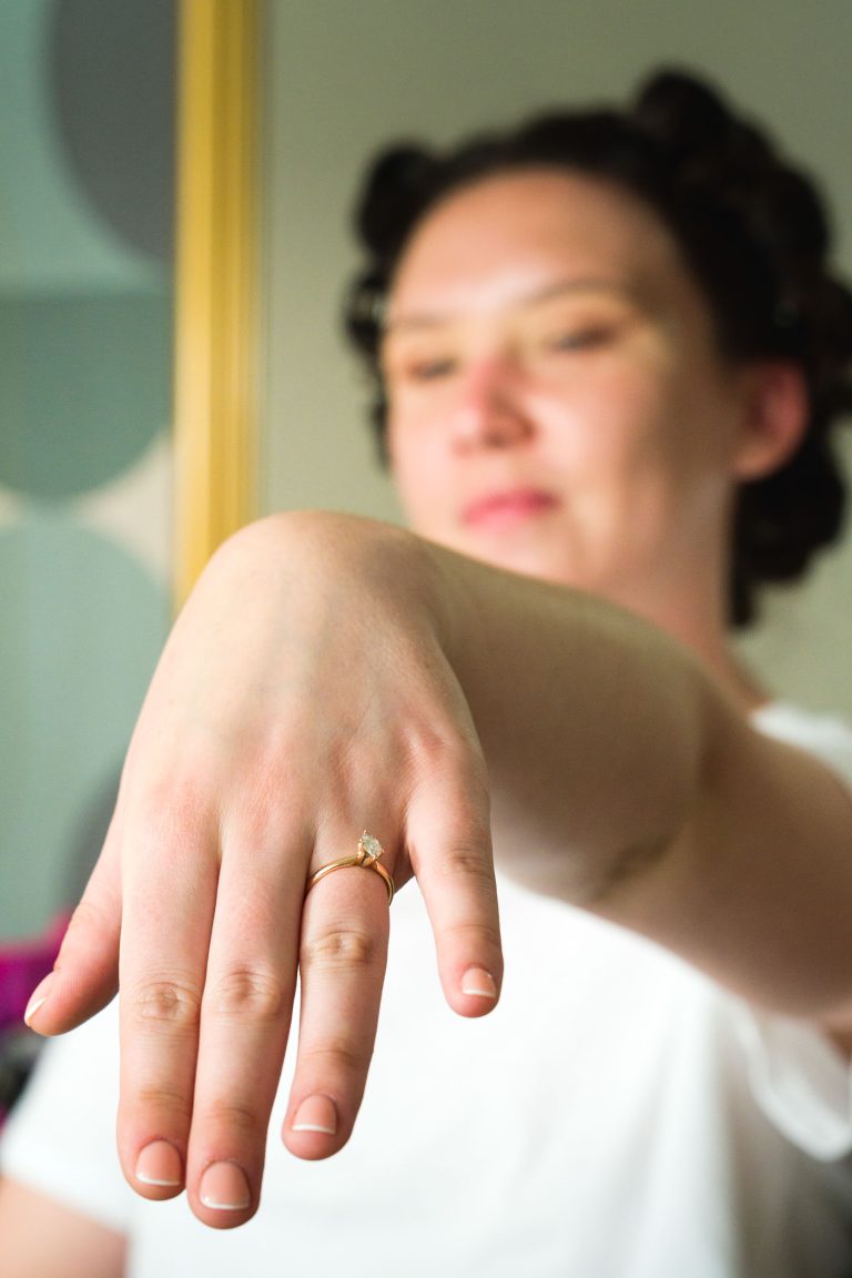 Une personne à la peau claire et aux cheveux foncés coiffés en rouleaux montre sa main gauche près de l'appareil photo, portant une bague en or avec un petit diamant. La mise au point se fait sur la main et la bague, tandis que le visage de la personne et l'arrière-plan sont flous.