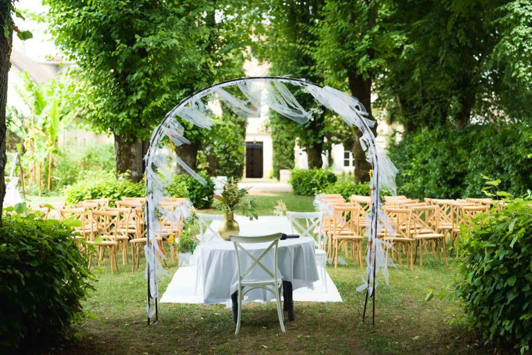 Un mariage en plein air est organisé autour d'une arche blanche drapée d'un tissu transparent, d'une table sous laquelle se trouvent un vase et une chaise, et de rangées de chaises en bois pour les invités. Des arbres et des buissons verdoyants entourent la scène, créant une atmosphère paisible et naturelle.