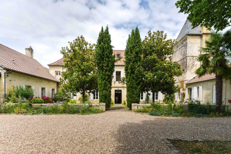 Un grand château en pierre avec de hautes fenêtres et un toit en tuiles se dresse derrière une cour gravillonnée, flanquée de jardins luxuriants, de grands cyprès, de buissons fleuris et d'arbres feuillus. L'entrée se compose d'une porte en bois encadrée de verdure sous un ciel partiellement nuageux.