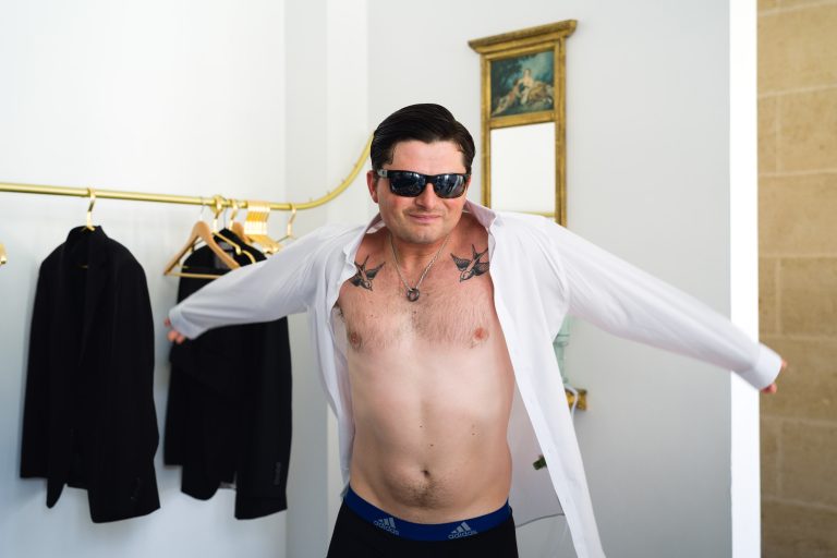 Un homme aux cheveux noirs et aux lunettes de soleil se tient à l'intérieur et enfile une chemise blanche. Il est torse nu en dessous, révélant des tatouages d'oiseaux sur sa poitrine. Des costumes sont suspendus à une étagère dorée derrière lui, et un miroir avec des œuvres d'art est accroché au mur blanc.