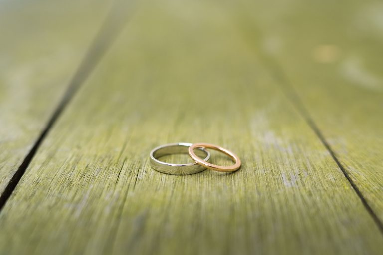 Deux anneaux, l'un en argent et l'autre en or, reposent ensemble sur une surface en bois vert et usé par les intempéries. Les anneaux sont simples et unis, positionnés de manière rapprochée, symbolisant le lien ou le partenariat. L'arrière-plan est légèrement flou, ce qui permet de concentrer l'attention sur les anneaux.