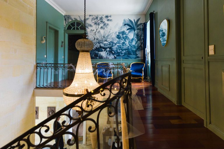 Un grand couloir vintage avec des murs verts, une rampe en fer et un grand lustre en cristal. Une peinture murale représentant des plantes tropicales est visible au-dessus de deux chaises bleues et d'une petite table. La lumière du soleil pénètre à flots, mettant en valeur le décor élégant et classique.