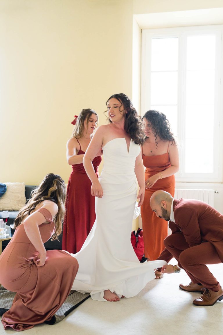 Une mariée vêtue d'une robe blanche sans bretelles se tient à l'intérieur, pieds nus, tandis que trois demoiselles d'honneur vêtues de robes couleur rouille et un homme en costume l'aident à ajuster sa robe. La lumière du soleil traverse une grande fenêtre derrière eux, créant une atmosphère chaleureuse et joyeuse.