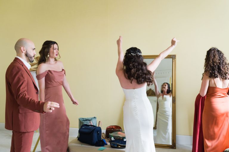 Dans une pièce aux murs jaune pâle, quatre personnes font la fête. Une femme en robe blanche regarde au loin, les bras levés. Deux femmes en robe cuivre et rouille et un homme barbu en costume la regardent en souriant. Un grand miroir et des sacs sont posés sur le sol à l'arrière-plan.