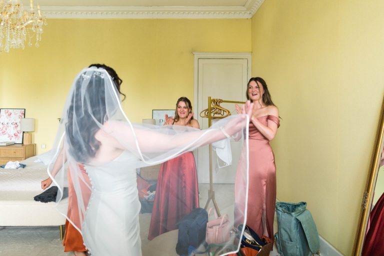 Une mariée vêtue d'une robe blanche et d'un voile virevolte joyeusement dans une pièce lumineuse, sous le regard de deux demoiselles d'honneur vêtues de robes rose pâle, qui sourient et applaudissent. La pièce est doucement éclairée, avec un lit défait, des vêtements et des accessoires visibles à l'arrière-plan.