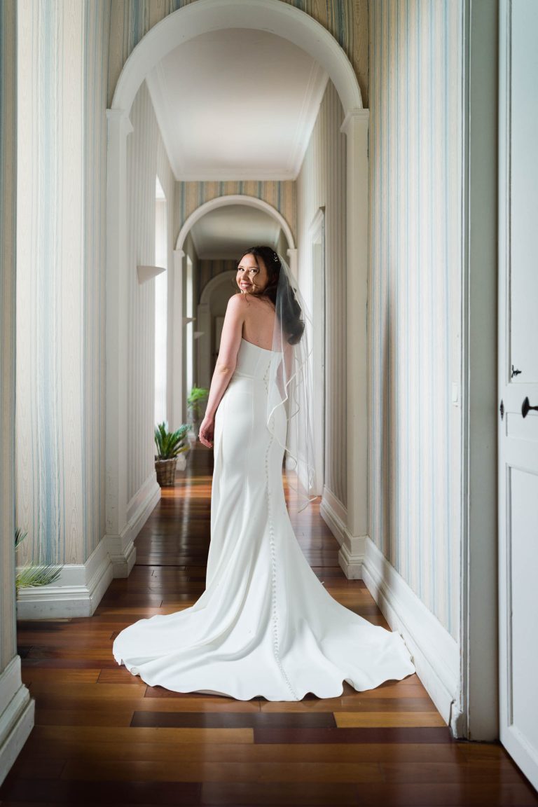 Une mariée se tient dans un couloir au sol en bois et au papier peint rayé. Elle fait face à la foule mais se retourne pour sourire par-dessus son épaule. Elle porte une robe blanche ajustée avec une longue traîne et un voile. La lumière des fenêtres éclaire le couloir et des plantes en pot tapissent les murs.