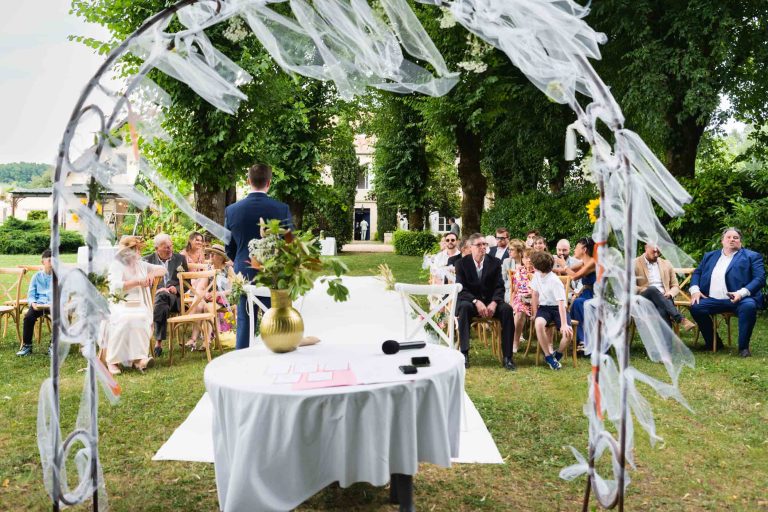 Cérémonie de mariage en plein air : les invités sont assis sur des chaises en bois face à une arche décorée de rubans blancs. Un homme se tient à l'avant, vu de dos, avec une table devant laquelle se trouvent un vase, des papiers et un microphone. Des arbres donnent de l'ombre à l'arrière-plan.