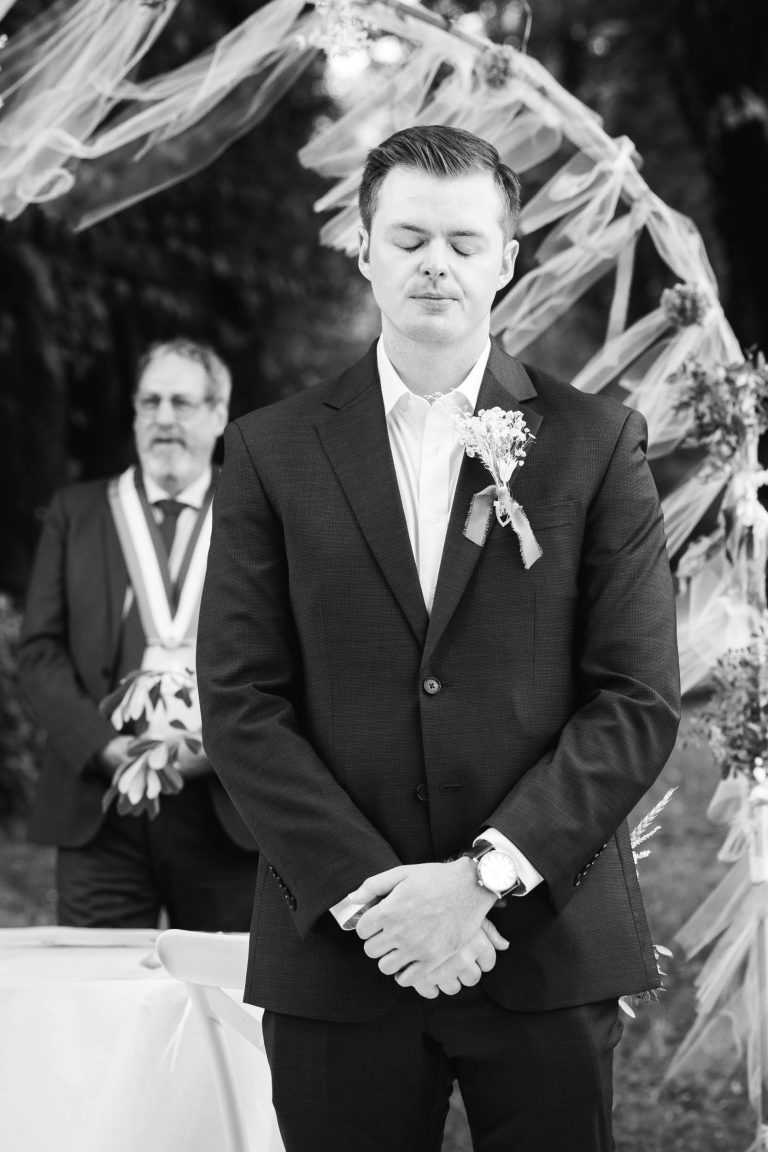 Un homme en costume se tient debout, les yeux fermés et les mains jointes, l'air solennel ou réfléchi, sous une arche décorée d'un tissu fluide. Un autre homme en costume se tient derrière lui. La scène suggère une cérémonie formelle, peut-être un mariage, en plein air.