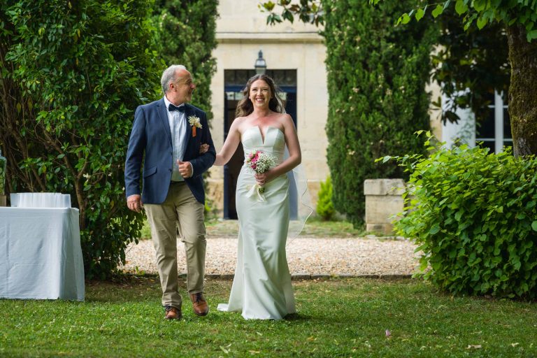 Une mariée souriante, vêtue d'une robe blanche sans bretelles, tient un bouquet et marche bras dessus bras dessous avec un homme plus âgé, vêtu d'un blazer bleu marine et d'un pantalon beige. Ils sont en plein air sur de l'herbe, entourés de verdure, avec un bâtiment en pierre à l'arrière-plan.