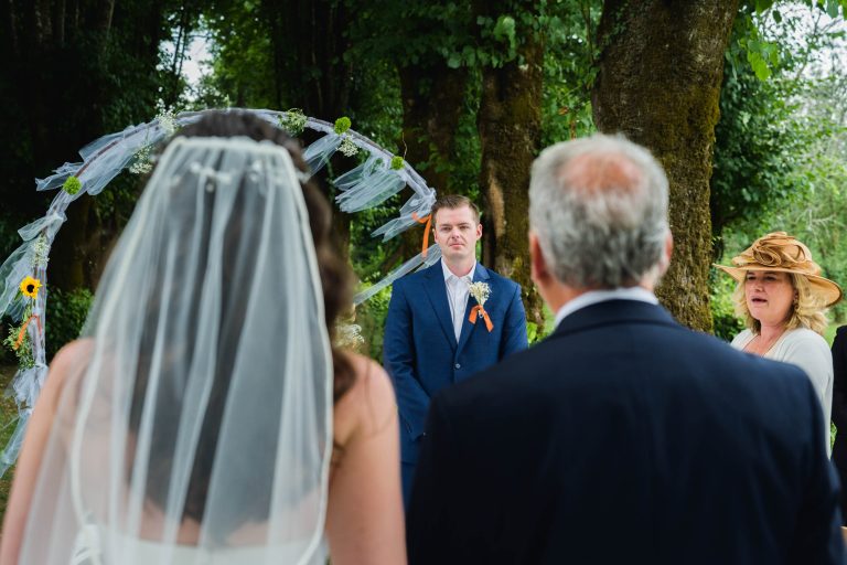 Une mariée vêtue d'une robe blanche et d'un voile se tient avec un homme plus âgé face à un marié vêtu d'un costume bleu, en plein air. Des arbres et une arche florale se trouvent à l'arrière-plan. Une femme plus âgée coiffée d'un chapeau beige regarde la scène depuis la droite. Tous semblent assister à une cérémonie de mariage.