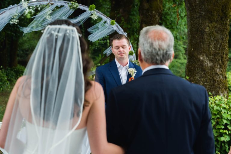 Une mariée vêtue d'une robe blanche et d'un voile se tient à côté d'un homme plus âgé, tous deux faisant face à un marié vêtu d'un costume bleu. Le marié se tient sous une arche décorée en plein air, avec des arbres feuillus en arrière-plan, avec un regard ému lors d'une cérémonie de mariage.